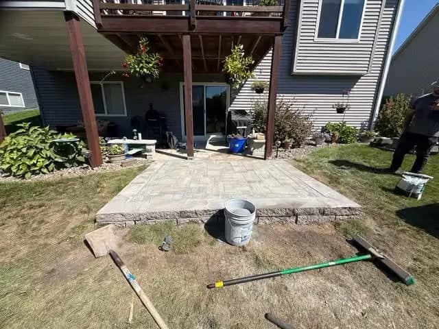 Patio installation in backyard with stone pavers and landscaping. Tools and materials visible.
