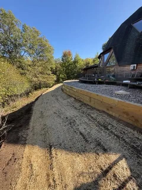 Graveled landscaping with straw near a wooden house in a wooded area under clear blue skies.